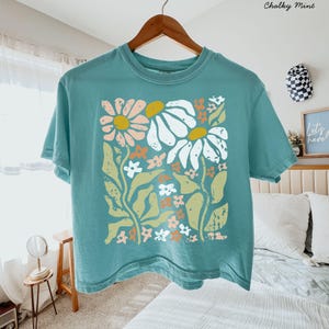 May include: A teal short-sleeve t-shirt featuring a large graphic print of stylized flowers in shades of white, light pink, mustard yellow, and sage green. The floral design has a distressed, textured appearance and is centered on the front.
