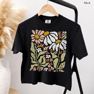 May include: A black short-sleeve t-shirt featuring a large, distressed graphic print of stylized flowers in shades of muted pink, white, mustard yellow, and sage green. The design includes two prominent daisies and smaller floral accents.