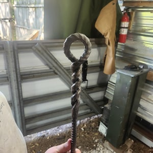 May include: A hand-forged metal tool with a twisted shaft and a circular loop at the top. The metal has a dark, textured finish, and the tool appears to be handmade. The background includes a workshop setting with a glimpse of an American flag.