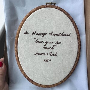 May include: An oval embroidery hoop with a faux wood grain frame containing off-white fabric. Dark brown thread is stitched into the fabric to display the text: Be Happy Sweetheart. love you so much, Mum & Dad, and a small heart symbol at the bottom.
