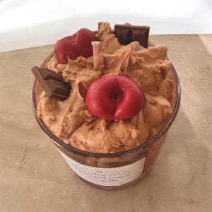 May include: A candle in a glass jar topped with brown wax textured like whipped frosting, decorated with two red cherry-shaped wax pieces and two small brown chocolate bar-shaped wax pieces. A label on the jar reads Country Pour Chocolate Cherry.