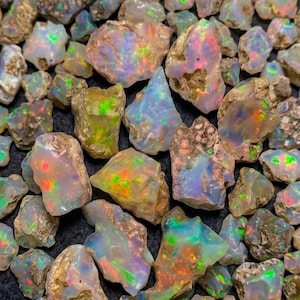 Pode incluir: Uma coleção de pedras de opala brutas e não polidas com uma base branca leitosa e flashes vibrantes de verde neon, laranja e azul. Cada pedra apresenta bordas irregulares e texturas naturais e ásperas em todo o grupo.
