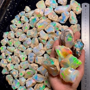 May include: A large collection of raw, unpolished opal stones featuring iridescent flashes of green, blue, orange, and yellow. A metal ruler is positioned on the right side, showing measurements in centimeters, with the stones spanning up to 30 cm.