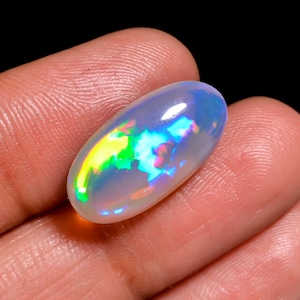 May include: An oval-shaped cabochon opal gemstone featuring a translucent, milky white base with vibrant flashes of iridescent green, yellow, and blue color play across its smooth, polished surface, reflecting light with a bright, glowing sheen.