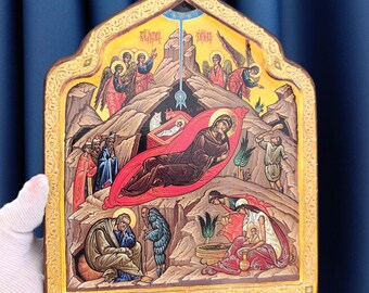Può includere: Un'icona religiosa in legno raffigurante una scena della Natività con una figura centrale in veste rossa, una mangiatoia con un neonato, angeli e varie figure su un paesaggio roccioso. L'icona ha una parte superiore appuntita e un bordo dorato.