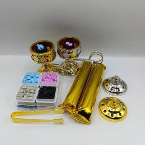 May include: Two gold-colored metal incense burners with filigree patterns, a set of small square containers holding blue, pink, white, and black incense pieces, a gold-toned pair of tongs, two gold-wrapped packages, and two circular lids.