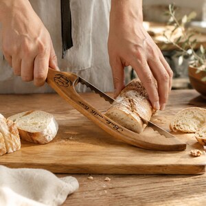 May include: A wooden bread slicer guide with a serrated metal blade, featuring a laser-engraved chef hat icon and the text "Bake with love" on the handle, used to slice a crusty loaf of bread on a wooden cutting board.