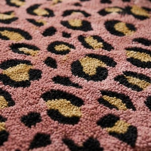 Puede incluir: Una alfombra de tufting con un patrón de leopardo repetitivo en tonos rosa empolvado, beige y negro. La superficie tiene una construcción de pelo grueso y texturizado con bucles suaves y distintos que crean un aspecto táctil en relieve.
