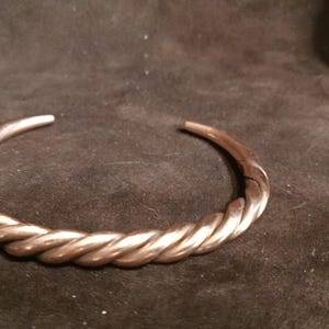Puede incluir: Una pulsera de brazalete metálico que presenta un diseño de cuerda trenzada continua. La pulsera está abierta en la parte posterior, formando una forma circular con extremos cónicos. La superficie tiene un acabado pulido y reflectante.