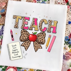 Peut inclure: Un sac fourre-tout blanc avec le mot TEACH en lettres imprimé léopard et rose. En dessous se trouve un grand nœud léopard avec une pomme rouge, un crayon rose, un crayon jaune, une règle rose et un carnet avec le texte Teach Love Inspire Motivate Lead Encourage.