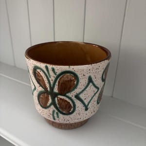 May include: A ceramic planter with a speckled cream-coloured exterior featuring hand-painted dark green diamond shapes and brown floral motifs. The interior is a solid brown glaze, and the base is a darker brown, providing a sturdy foundation.