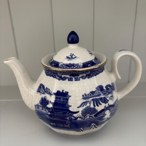 May include: A white ceramic teapot featuring a blue willow pattern, a matching lid with a blue finial, and a gold-trimmed rim. The teapot body displays a detailed landscape illustration with pagodas, trees, and bridges, finished with a fine crackle glaze texture throughout the surface.
