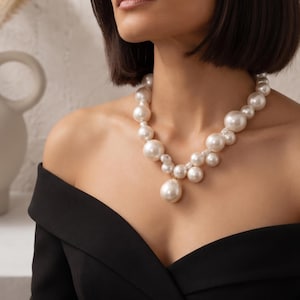 May include: A statement necklace featuring a collection of white, spherical pearls of varying sizes arranged in a cluster. The design includes a prominent, larger teardrop-shaped pearl pendant hanging from the center of the beaded strand.