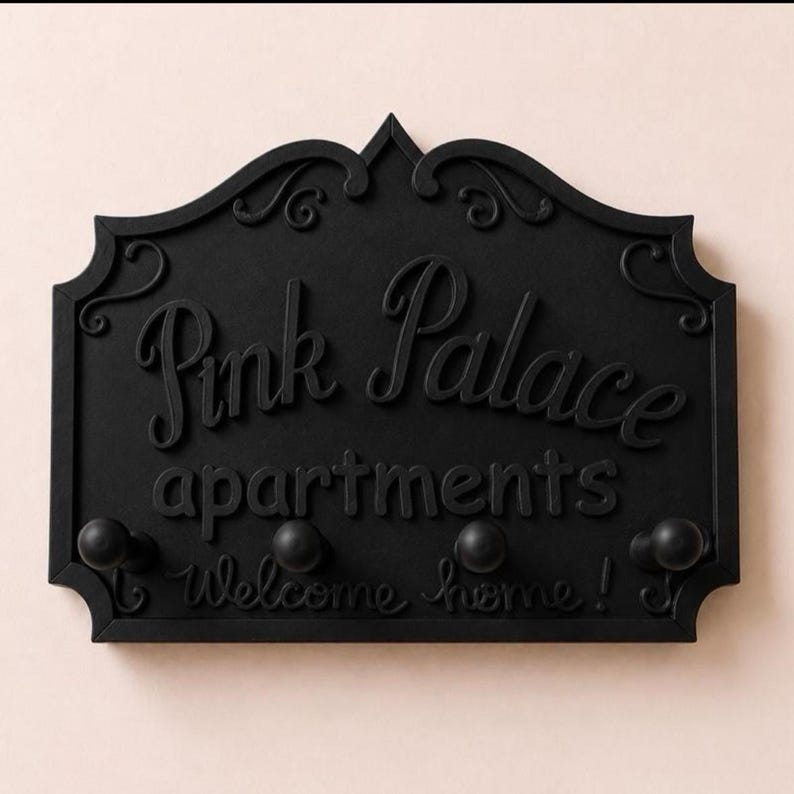 Może przedstawiać: Czarny znak ścienny z napisem "Pink Palace apartments Welcome home!" w ozdobnej czcionce. Znak ma cztery okrągłe haczyki pod tekstem i ozdobne zawijasy na g&oacute;rze.