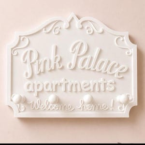 Może przedstawiać: Biały dekoracyjny szyld ścienny z napisem "Pink Palace apartments" w ozdobnej czcionce. Znak ma dekoracyjną obw&oacute;dkę i zawiera frazę "Welcome home!" pod nazwą apartamentu. Zawiera trzy małe gałki.