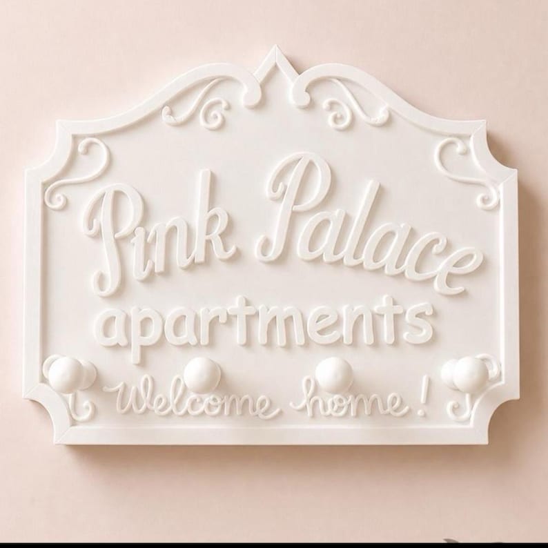 Może przedstawiać: Biały dekoracyjny szyld ścienny z napisem "Pink Palace apartments" w ozdobnej czcionce. Znak ma dekoracyjną obw&oacute;dkę i zawiera frazę "Welcome home!" pod nazwą apartamentu. Zawiera trzy małe gałki.