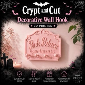 Może przedstawiać: R&oacute;żowy, drukowany w 3D dekoracyjny wieszak ścienny z napisem "Pink Palace apartments welcome home!". Wieszak ma dekoracyjną obw&oacute;dkę i jest zaprojektowany w stylu Halloween. Obraz zawiera r&oacute;wnież nietoperze, nagrobki i tekst "Crypt and Cut".