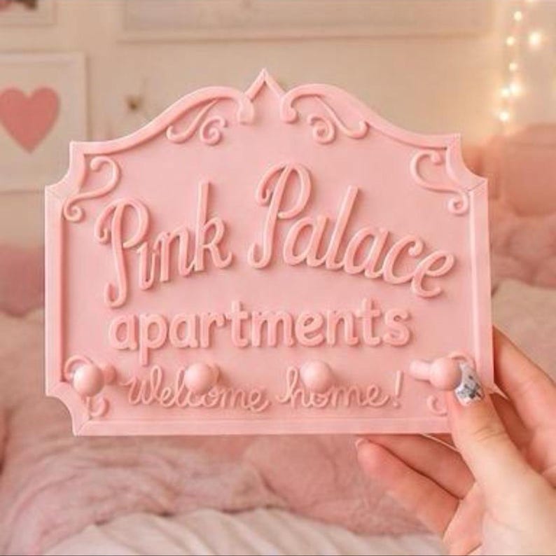Może przedstawiać: R&oacute;żowy znak ścienny z napisem "Pink Palace apartments welcome home!" w ozdobnej czcionce. Znak ma falisty brzeg i trzy małe gałki do wieszania przedmiot&oacute;w. Tło jest w kolorze miękkiego r&oacute;żu.