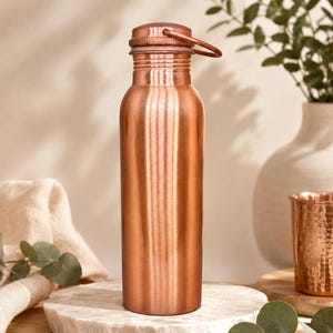 May include: A cylindrical copper water bottle with a smooth, polished finish and a matching screw-top lid featuring a small integrated carrying loop. The bottle has a narrow neck and a wide base, reflecting light across its metallic surface.