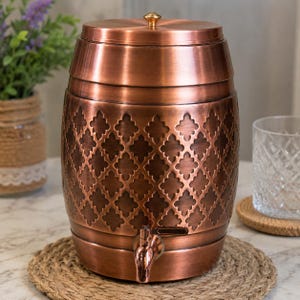 May include: A cylindrical copper beverage dispenser with a barrel-like shape, featuring a repeating quatrefoil pattern embossed on the exterior. It includes a matching copper lid with a small knob and a functional spigot at the base.