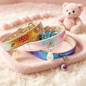 May include: Three pet collars with a holographic, iridescent scale pattern. The collars feature a metallic finish in rainbow, light blue, and purple tones. Each collar includes a silver-toned buckle, a metal D-ring, and a dangling charm pendant.