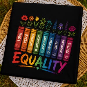 May include: A black t-shirt featuring a graphic of nine colourful book spines arranged in a row. Each book is labelled with words including Love, Peace, Inclusion, Kindness, Acceptance, Diversity, Unity, Compassion, and Justice. Above the books are various flowers, and the word Equality is printed in large, rainbow-coloured letters at the bottom.
