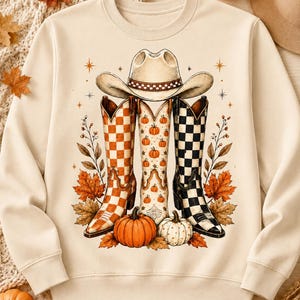 Puede incluir: Una sudadera de cuello redondo color crema con un estampado gráfico de un sombrero de vaquero sobre tres botas vaqueras. Las botas muestran diseños de cuadros naranjas y blancos, estampados de calabazas y cuadros negros y blancos con hojas.