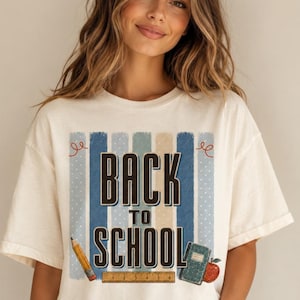Peut inclure: Un t-shirt à manches courtes blanc cassé avec un imprimé graphique à rayures verticales bleues et beiges. Le texte BACK TO SCHOOL est écrit en lettres sombres. Sous le texte se trouvent une règle en bois, un crayon jaune, un livre vert et une pomme rouge.