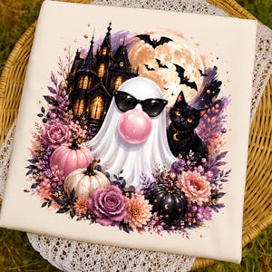 May include: A white fabric square featuring a graphic print of a ghost wearing black sunglasses and blowing a pink bubble, a black cat, a dark mansion, a full moon with bats, and a decorative arrangement of pink, white, and black pumpkins and flowers.