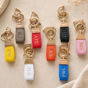 Puede incluir: Un conjunto de ocho llaveros rectangulares de cuero en varios colores, incluyendo amarillo, gris, rojo, naranja, blanco, negro, azul y rosa. Cada llavero cuenta con una anilla de metal dorado y una tapa dorada con iniciales grabadas.