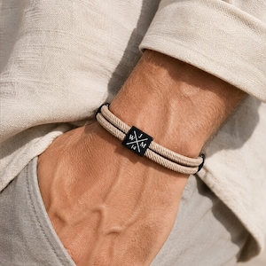 Puede incluir: Una pulsera de doble cordón beige con una pieza central cuadrada de metal negro. La pieza central está grabada con un diseño de cruz blanca, las letras W, J y M, y el número 14. La pulsera tiene terminales de metal negro.