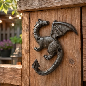 Può includere: Un gancio da parete in ghisa nera a forma di drago montato su un palo di legno verticale. La figura del drago è raffigurata di profilo con le ali spiegate, una coda curva che termina con una punta e due fori per le viti di fissaggio.