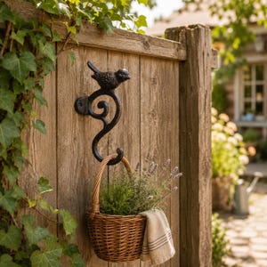 May include: A black cast iron wall hook featuring a bird figure perched atop a decorative scroll design. A woven wicker basket containing green plants and a small beige cloth with thin stripes hangs from the bottom curve of the metal hook.
