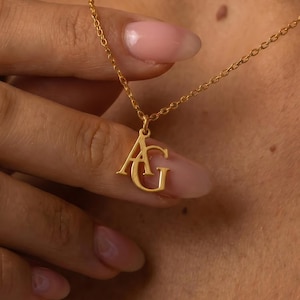 May include: A gold-colored necklace with a delicate chain. The pendant features the intertwined letters "AG". The necklace is displayed against a skin-toned background.