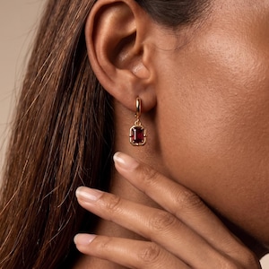 May include: A gold hoop earring featuring a rectangular, deep red gemstone pendant. The stone is set in a polished gold frame with rounded corners, suspended from a small, circular gold hoop that secures through the earlobe.