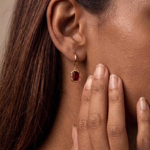 May include: A gold hoop earring featuring a rectangular, deep red gemstone pendant. The stone is set in a decorative gold frame with small bead-like accents along the edges. The earring hangs from a simple, polished gold hoop that secures to the ear.