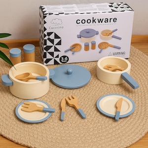 Peut inclure: Un ensemble de jouets de cuisine en bois avec une boîte indiquant "cookware". L'ensemble comprend des casseroles, des poêles, des ustensiles, des assiettes et des salières. La batterie de cuisine est de couleur crème avec des accents bleus.