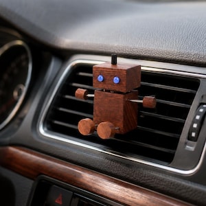 Op de afbeelding: Een kleine, bruine, robotvormige auto-luchtverfrisserclip bevestigd aan een zwart ventilatierooster van het dashboard. De robot heeft een rechthoekig hoofd en lichaam, twee kleine blauwe ronde ogen en cilindrische houten armen en benen.