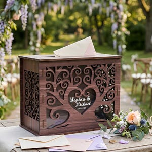 May include: A rectangular dark brown wooden card box with intricate laser-cut scrollwork and a central heart cutout. The front panel features the white printed text "Sophia & Mickael" and "08/15/56". A light-coloured envelope is inserted into the top slot.