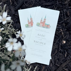 May include: Two wedding ceremony programs with a watercolor illustration of a church. The programs are light blue with text that reads "Alec and Nicole" and the wedding date. The programs are set against a dark background with white flowers.