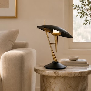 May include: A table lamp featuring a matte black saucer-shaped shade and a brass-toned metal stand. The lamp has a central brass socket holding a single light bulb, supported by an angled brass arm connected to a circular black base.