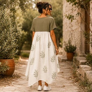 May include: A midi-length dress featuring a solid olive green short-sleeved bodice and a cream-colored skirt. The skirt is decorated with a repeating pattern of olive green leaf sprigs. The dress has a loose, flowing silhouette and a round neckline.