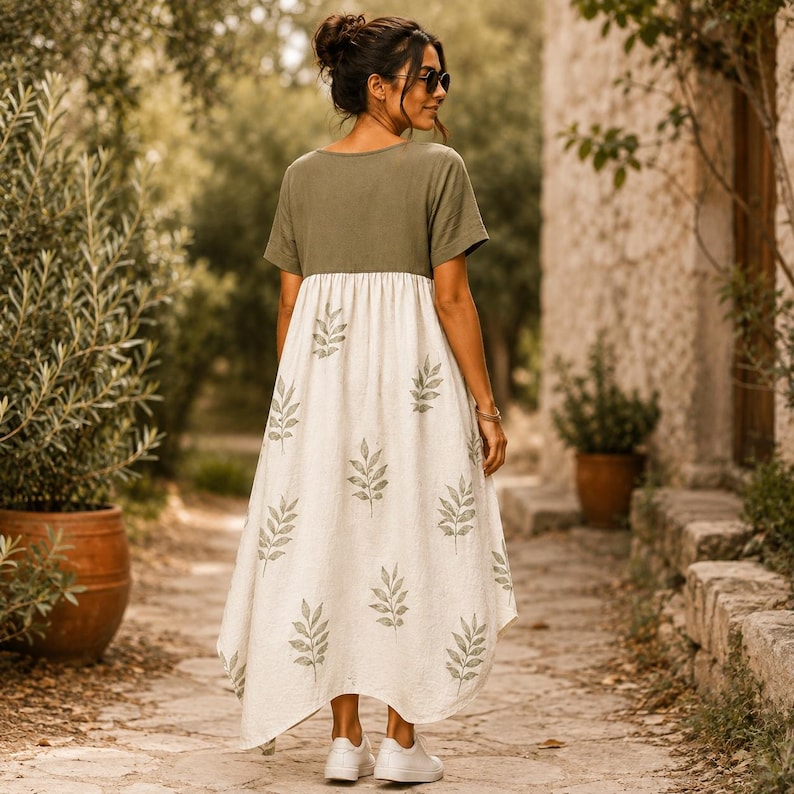 May include: A midi-length dress featuring a solid olive green short-sleeved bodice and a cream-colored skirt. The skirt is decorated with a repeating pattern of olive green leaf sprigs. The dress has a loose, flowing silhouette and a round neckline.