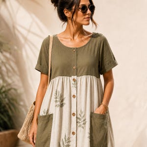 May include: A two-tone dress featuring an olive green bodice and a cream-colored skirt with a green leaf print. The dress has short sleeves with cuffs, a round neckline, a central button-down placket, and two large olive green patch pockets.