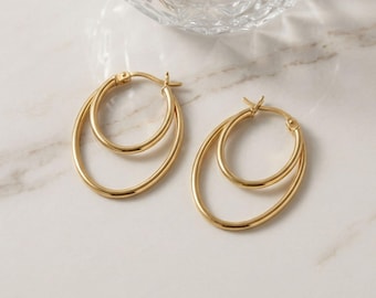 May include: A pair of gold-coloured hoop earrings. Each earring has a double oval design, with a smaller hoop inside a larger one. The earrings are displayed on a white marble surface, reflecting light.