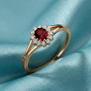 May include: A gold ring featuring a central oval, deep red gemstone encircled by small, clear gemstones. The ring's band splits into two delicate strands. The ring is set against a blue background.