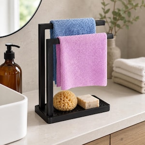 Può includere: Un portasciugamani in metallo nero con due barre orizzontali che sostengono un asciugamano blu e uno rosa. La base presenta un vassoio perforato che ospita una spugna naturale rotonda e una saponetta rettangolare.