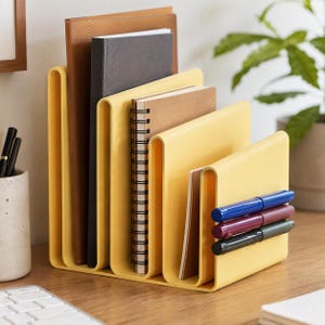 May include: A yellow plastic desk organizer with four vertical slots holding various notebooks and folders. The front section features three horizontal pen holders containing one blue, one maroon, and one black pen, keeping stationery items organized.