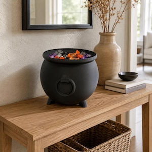 Puede incluir: Un caldero negro mate con cuerpo redondeado, tres pies pequeños y un asa de anillo decorativa en la parte frontal. El interior está lleno de una colección de gemas de plástico translúcidas, multicolores y de formas variadas en tonos naranja y morado.