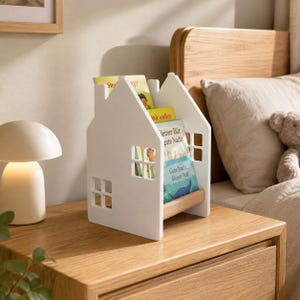 Könnte beinhalten: Ein weißes, hausförmiges Bücherregal aus Holz mit ausgeschnittenen Fensterdetails an den Seiten. Das Gestell enthält mehrere Kinderbücher, darunter eines mit dem sichtbaren Text "Kleiner Bär gute Nacht" und "Gute Nacht, kleiner Wal" auf dem Einband.